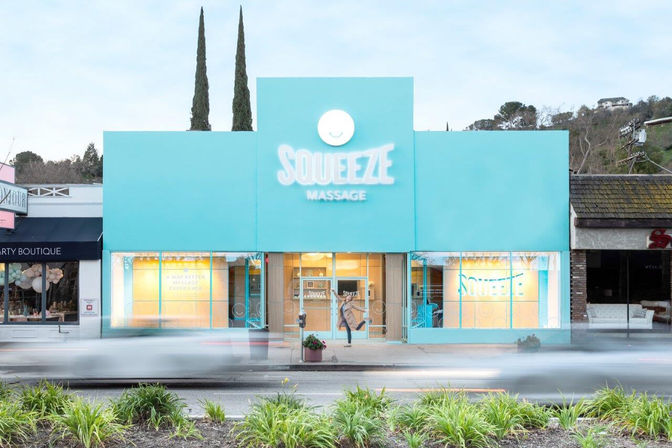 Bright turquoise massage spa storefront on a busy main street, warm-lit windows, blurred cars passing, a person entering, tall cypress trees and neighboring shops visible