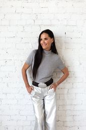 Smiling woman with long dark hair wearing a gray short-sleeve top, wide black belt and metallic silver pants, hands in pockets, posing against a white brick wall — chic minimalist fashion portrait.