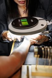 Close-up of a tattooed nail technician shaping a client's nails under a bright magnifying lamp in a nail salon, rings and manicure tools visible.