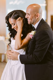 Wedding couple embracing at an indoor reception — close-up of bride in white gown and groom in black tux with pink boutonniere, sharing a joyful, intimate moment.