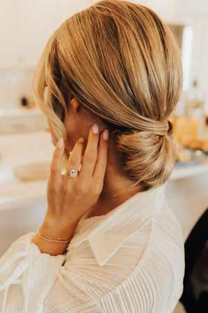 Close-up of a blonde low-bun updo with an elegant twist, hand showing a sparkling engagement ring and gold bracelets, white textured blouse in a soft bridal-prep setting.