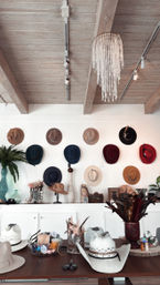 Rustic boutique hat shop interior with exposed wooden beams and a crystal chandelier, wall display of colorful wide-brim and felt hats, and a table filled with cowboy hats, feathers and hat-making supplies.