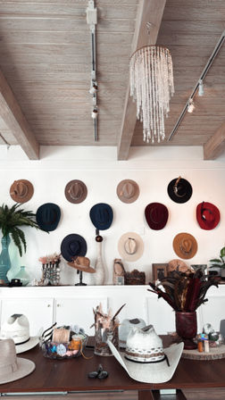 Rustic boutique hat shop interior with exposed wooden beams and a crystal chandelier, wall display of colorful wide-brim and felt hats, and a table filled with cowboy hats, feathers and hat-making supplies.