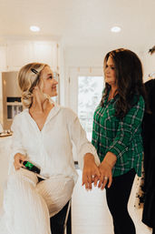 Cheerful getting-ready scene in a bright kitchen: woman in white loungewear sits holding a green trimmer while a friend in a green plaid shirt stands, they smile and hold hands showing rings.