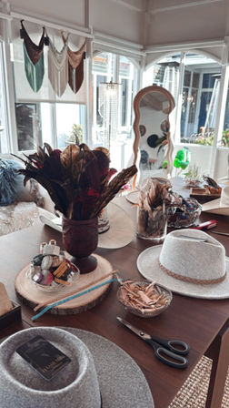 Sunlit boho hat workshop table with gray felt fedoras, decorative feather arrangements, hat trims and beaded bands on a wooden slab, scissors and a wavy mirror in a bright studio.
