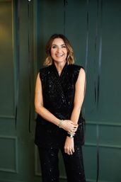 Smiling woman in a black sequined suit with wavy bob hair posing indoors against a dark green paneled wall with hanging black streamers.