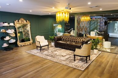 Stylish boutique lounge with dark leather Chesterfield sofa, two cream armchairs, ornate gold mirror, hat display rack, green paneled walls, golden chandelier and patterned rug on wood floors