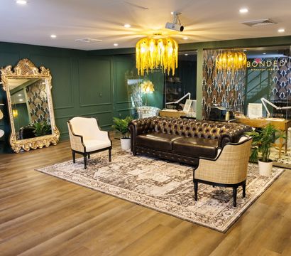 Stylish boutique lounge with dark leather Chesterfield sofa, two cream armchairs, ornate gold mirror, hat display rack, green paneled walls, golden chandelier and patterned rug on wood floors