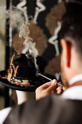 Person seen from behind directs a metal tube that emits curling smoke onto a black felt hat with a worn patterned band displayed on a mannequin head.