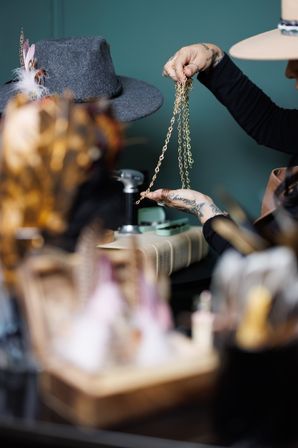 Tattooed hands in an artisan studio holding dangling gold chains above a gray wool fedora with feather accents on a cluttered workbench