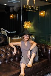 Stylish woman in a black hat and neutral vest-and-pants outfit lounging on a tufted brown leather sofa in a chic boutique jewelry studio, wearing multiple rings, bracelets and an anklet with warm green walls and soft lighting.