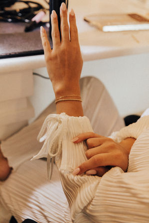 Close-up of hands and forearm resting on a table, pleated white blouse sleeve with tied cuff, delicate gold bracelets and pale pink manicure — cozy indoor lifestyle beauty detail.
