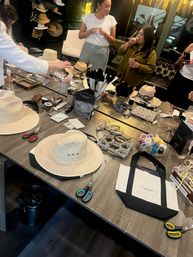 Bustling hat-decorating workshop in a modern studio — communal table filled with straw hats, ribbons, feathers, spools, scissors and craft supplies while participants create.