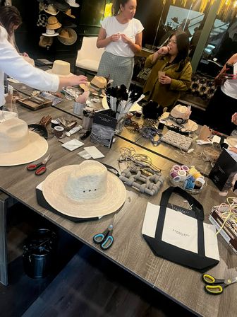 Bustling hat-decorating workshop in a modern studio — communal table filled with straw hats, ribbons, feathers, spools, scissors and craft supplies while participants create.