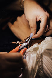 Close-up of hands using small pliers to repair a delicate gold chain on a leather band, blue-painted nails and textured fabric in warm workshop lighting.