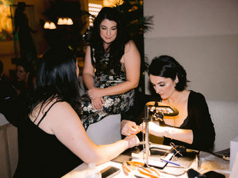 Three women at an indoor evening event — a nail technician works on a guest’s manicure under a bright desk lamp while another woman in a sequined dress watches.