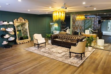 Upscale boutique salon lounge with tufted dark brown leather sofa, two cream armchairs on a patterned rug, ornate gold-framed mirror, hat display, green paneled wall, dripping-gold chandelier and glass partition leading to a stylish studio