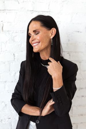 Smiling woman with long dark hair and closed eyes wearing a black blazer and silver jewelry, posing against a white brick wall in a stylish studio portrait.