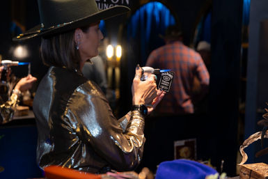 Woman in a wide-brimmed hat and shiny metallic jacket holding up a small Jack Daniel’s tin in a dim, blue-lit lounge with blurred patrons in the background
