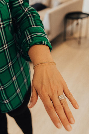 Close-up of a hand wearing two delicate gold bracelets and a sparkling diamond ring, cuff of a green plaid shirt visible against a blurred indoor background.