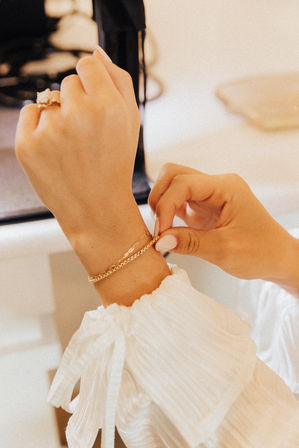 Close-up of hands fastening a gold chain bracelet on a wrist over a white ruffled blouse with polished nails