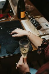 Close-up of hands wearing delicate bracelets and rings beside a rotating bracelet display and champagne glass on a boutique wooden table at an artisan jewelry pop-up