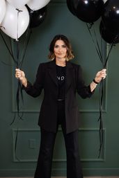 Stylish woman in a black blazer and pants holding clusters of black and white balloons against a green paneled wall — modern fashion portrait with a celebratory vibe.