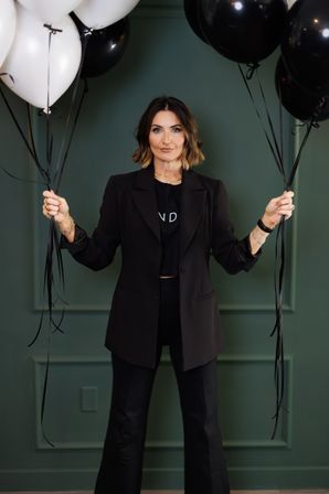 Stylish woman in a black blazer and pants holding clusters of black and white balloons against a green paneled wall — modern fashion portrait with a celebratory vibe.