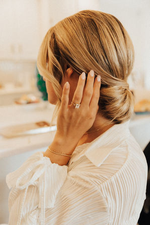 Close-up of a blonde low-bun hairstyle with a manicured hand showing a square-cut engagement ring and gold bracelets, wearing a white textured blouse in an elegant indoor getting-ready scene