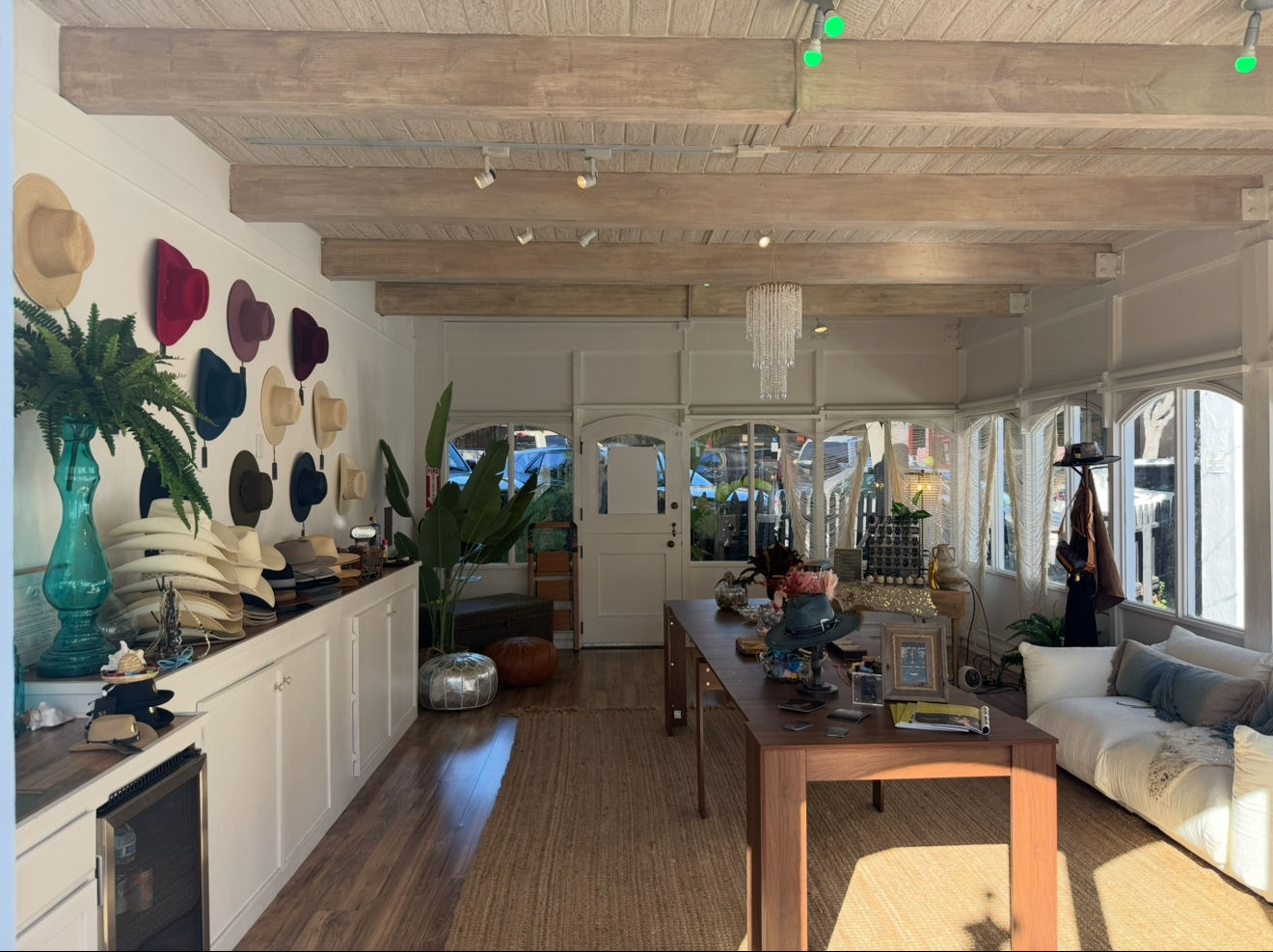 Sun-drenched boutique hat shop interior with colorful wide-brim hats on a white wall, stacked hat displays on a long counter, exposed wooden beams, central wooden table with accessories, plants and a cozy sofa by arched windows