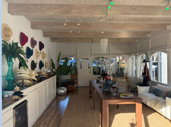 Sun-drenched boutique hat shop interior with colorful wide-brim hats on a white wall, stacked hat displays on a long counter, exposed wooden beams, central wooden table with accessories, plants and a cozy sofa by arched windows
