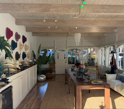 Sun-drenched boutique hat shop interior with colorful wide-brim hats on a white wall, stacked hat displays on a long counter, exposed wooden beams, central wooden table with accessories, plants and a cozy sofa by arched windows