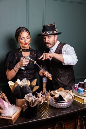 Two artisans at a millinery-style craft table, one using a small torch while the other guides a tool, surrounded by feathers, ribbons, spools and trims in a cozy studio setting.