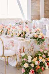 Elegant wedding reception table with floral tablecloth, lush pastel rose centerpieces spilling onto the floor, tall white taper candles, crystal glassware and gold-trimmed chairs in a bright ballroom