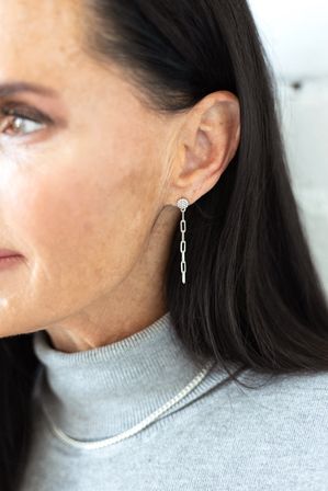 Close-up of a woman wearing sleek silver dangle chain earrings with a pavé round stud and linked rectangular drops, dark hair, gray turtleneck and delicate silver necklace