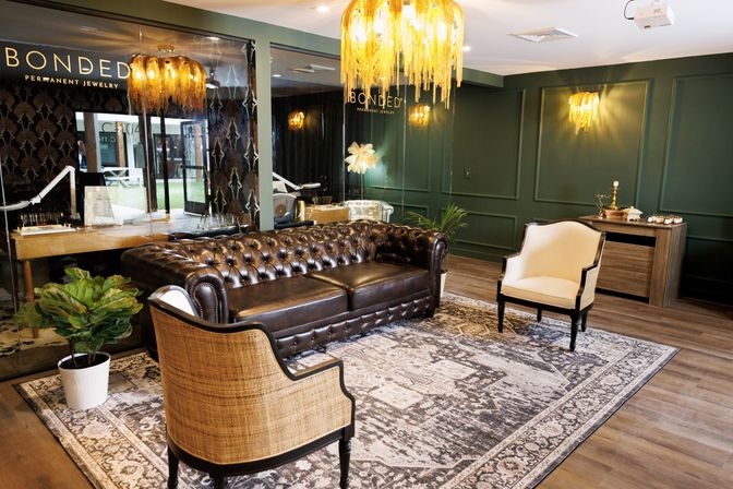 Upscale boutique waiting area with dark brown leather Chesterfield sofa, rattan and cream armchairs on a patterned rug, green paneled walls and gold fringe chandeliers.