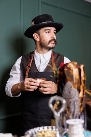 Bearded artisan in a black hat and leather‑strapped apron in a green-walled workshop, holding a small leather patch with craft tools blurred in the foreground.