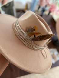 Close-up of a beige wide-brim felt fedora with layered ribbon band and mixed decorative feathers — boho-chic hat accessory.