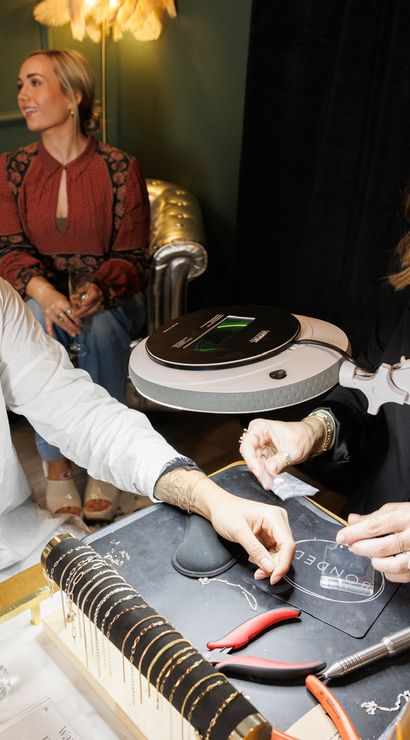 Smiling customer with a glass of champagne getting a bracelet fitted by a jeweler at a cozy boutique jewelry studio, tools and bracelet display on the table and a friend seated in the background.