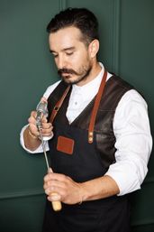 Bearded man in a leather‑trim apron using a handheld blowtorch to heat a metal skewer against a dark green background.