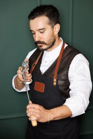 Bearded man in a leather‑trim apron using a handheld blowtorch to heat a metal skewer against a dark green background.