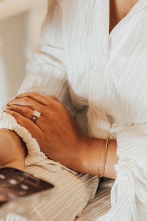 Close-up of hands resting on a white textured dress, featuring a square-cut diamond engagement ring, delicate gold bracelets and a smartphone at the edge in warm tones.
