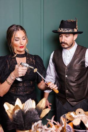 Two artisans in a craft studio — a woman in black lace uses a handheld torch to heat a small piece on a metal rod held by a man in a leather vest and feathered hat, with spools and decorative feathers on the table.