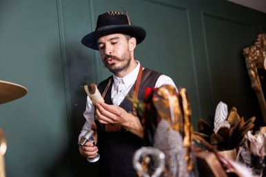 Artisan milliner in a black hat and leather apron using a handheld torch to shape a hat piece in a green-paneled workshop surrounded by hats and feathers