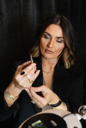 Woman with shoulder-length bob and layered necklaces examining a small jewelry piece with tweezers under a tabletop magnifying lamp, wearing multiple rings and bracelets against a dark backdrop.