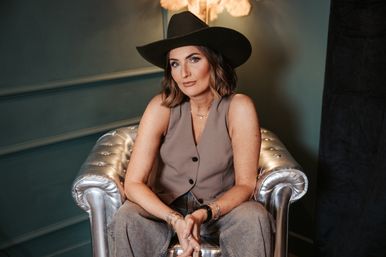 Stylish portrait of a woman in a black cowboy hat, sleeveless taupe vest and jeans seated on a silver tufted armchair in a moody teal indoor setting.