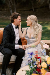 Bride in a lace jeweled gown and groom in a black tuxedo smiling on a vintage outdoor sofa amid colorful wedding florals on a grassy lawn