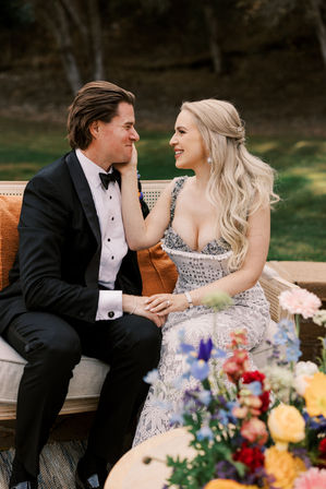 Bride in a lace jeweled gown and groom in a black tuxedo smiling on a vintage outdoor sofa amid colorful wedding florals on a grassy lawn