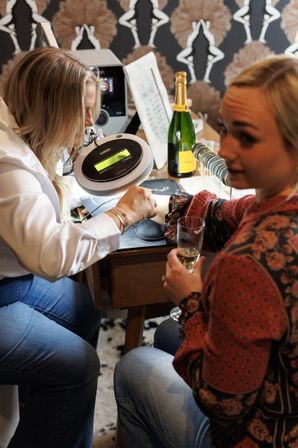 Woman receiving a manicure from a nail technician at a salon table — hands under a magnifying lamp with a champagne bottle and glass nearby and patterned wallpaper backdrop.