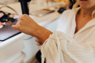Close-up of a person in a white tie-sleeve blouse wearing a delicate gold bracelet while using a black hair-styling tool at a vanity.
